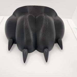 May include: A black, 3D-printed paw with four prominent claws. The paw has a rounded shape with a textured surface, and the claws are pointed. The object is set against a white background.