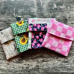 May include: Four fabric pouches with different patterns are displayed. The patterns include a map design, green gingham with sunflowers, a floral print with pink roses, and a pink and white checkerboard pattern. The pouches are folded over.