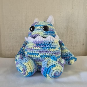 May include: A handmade crochet monster doll in shades of blue, purple, and yellow. The plush toy has large black eyes, small white horns, and a white frilly collar. The doll is sitting on a white surface.
