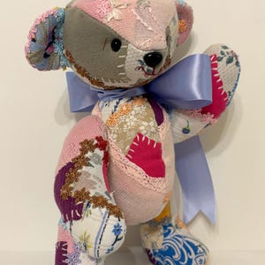 Puede incluir: Un oso de peluche patchwork hecho a mano con una cinta azul claro. El oso está hecho de varios retazos de tela en rosa, gris y blanco, con estampados florales y geométricos. La nariz y los ojos del oso son negros.