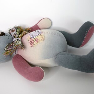 May include: A gray and pink patchwork teddy bear with a floral scarf lying on its back. The bear has a stitched seam around the edges of its limbs and body.
