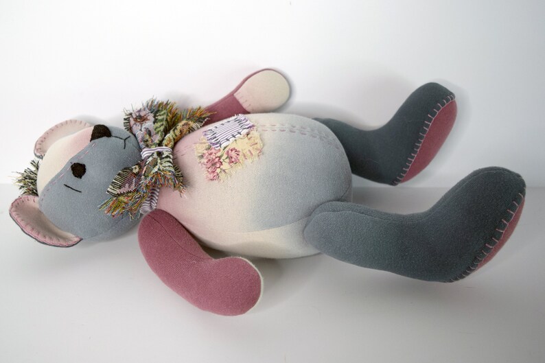 May include: A gray and pink patchwork teddy bear with a floral scarf lying on its back. The bear has a stitched seam around the edges of its limbs and body.