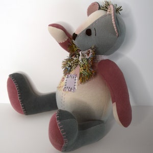 May include: A handmade patchwork teddy bear with gray and pink fabric. The bear has a colorful scarf around its neck and is sitting on a white surface.