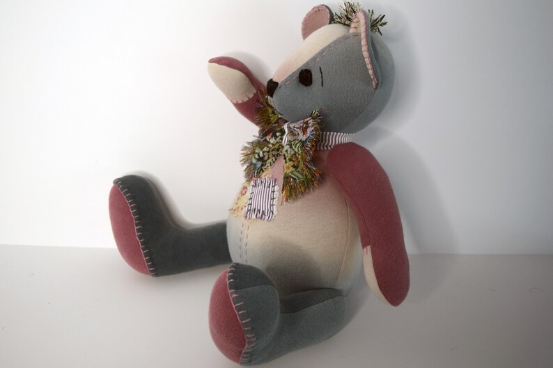 May include: A handmade patchwork teddy bear with gray and pink fabric. The bear has a colorful scarf around its neck and is sitting on a white surface.