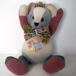 May include: A gray and pink patchwork teddy bear with a floral bow tie. The bear has a stitched smile and is sitting with its arms raised.