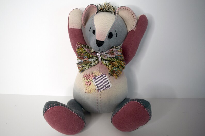 May include: A gray and pink patchwork teddy bear with a floral bow tie. The bear has a stitched smile and is sitting with its arms raised.