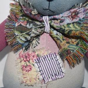 May include: A handmade patchwork teddy bear with a gray face, pink and gray fabric, and a colorful bow tie. The bear has a patch of fabric with flowers and stripes on its chest.