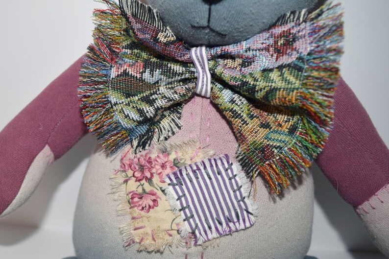 May include: A handmade patchwork teddy bear with a gray face, pink and gray fabric, and a colorful bow tie. The bear has a patch of fabric with flowers and stripes on its chest.