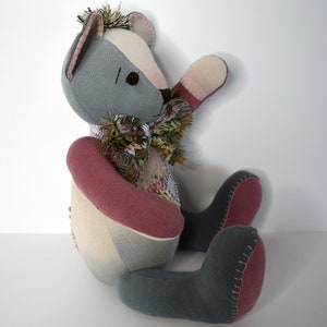 May include: A patchwork teddy bear with gray, pink, and white fabric. The bear is sitting with its right arm raised and its left arm resting on its lap. The bear has a scarf made of different fabrics around its neck.
