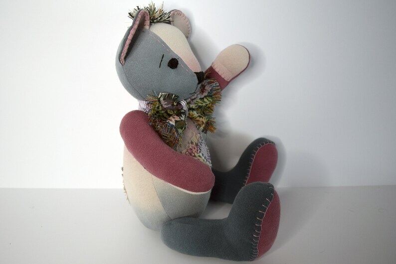 May include: A patchwork teddy bear with gray, pink, and white fabric. The bear is sitting with its right arm raised and its left arm resting on its lap. The bear has a scarf made of different fabrics around its neck.