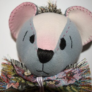 May include: A gray and pink handmade stuffed animal with a black nose and embroidered eyes. The animal is wearing a colorful, fringed scarf.