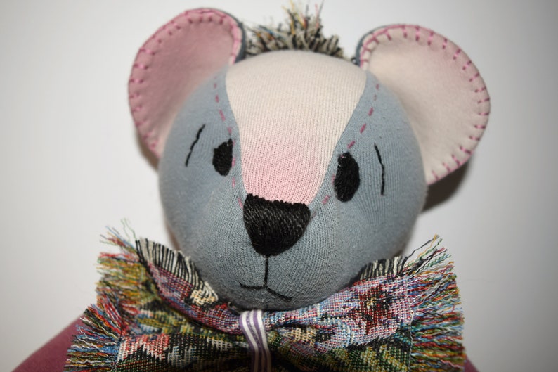 May include: A gray and pink handmade stuffed animal with a black nose and embroidered eyes. The animal is wearing a colorful, fringed scarf.