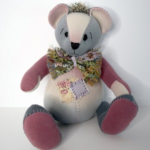 May include: A gray and pink patchwork teddy bear with a floral bow tie. The bear is sitting on a white surface.