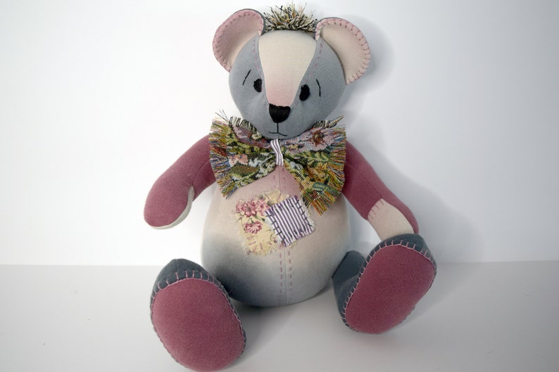 May include: A gray and pink patchwork teddy bear with a floral bow tie. The bear is sitting on a white surface.