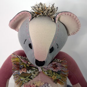 May include: A handmade plush toy bear with gray and pink fabric, a black nose, and a brown and green patterned bow tie.