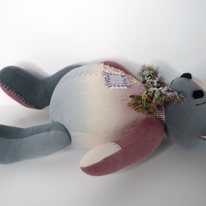 May include: A gray and pink patchwork teddy bear with a frayed fabric scarf lying on its back. The bear has black eyes and a stitched nose.