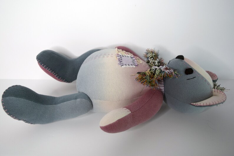May include: A gray and pink patchwork teddy bear with a frayed fabric scarf lying on its back. The bear has black eyes and a stitched nose.