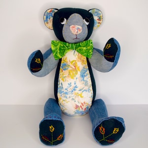 Puede incluir: Un oso de peluche de patchwork azul y blanco con una pajarita verde. El oso está sentado con las piernas extendidas y tiene flores bordadas en los pies.