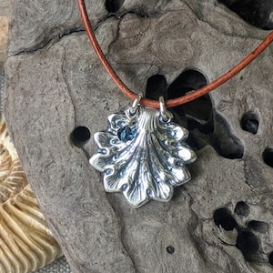 Può includere: Una collana con un ciondolo a forma di conchiglia di mare argentata con un accento di pietra preziosa blu su un cordoncino di cuoio marrone.