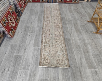 Op de afbeelding: Een lange, rechthoekige beige loper met een ingewikkeld patroon van bloemen en geometrische ontwerpen. De loper is gecentreerd op een grijze houten vloer, met verschillende rode en beige decoratieve tapijten en een rieten stoel op de achtergrond.