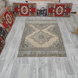 Puede incluir: Una alfombra rectangular beige con un intrincado diseño de medallón central en tonos grises, azules y marrones. La alfombra tiene un borde detallado y está colocada sobre un suelo de madera gris claro. Otras alfombras y cojines son visibles en el fondo.
