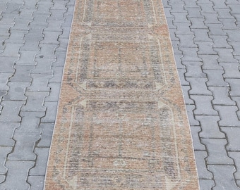 May include: A rectangular, distressed runner rug with a faded, vintage appearance. The rug features a central design in shades of brown, beige, and muted orange, with geometric patterns and borders. The rug is placed on a gray brick surface.