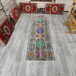 May include: A rectangular, vintage-style rug with a central pattern of green, purple, and orange floral motifs. The rug has a distressed look with a faded color palette, and measures approximately 3 feet wide.