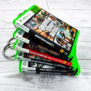 Può includere: Una pila di custodie di giochi per Xbox 360, tenute insieme da un anello d'argento. I giochi includono "Grand Theft Auto V", "Mortal Kombat", "Red Dead Redemption", "Grand Theft Auto: San Andreas" e "Call of Duty: Black Ops II". Le custodie hanno un bordo verde brillante.