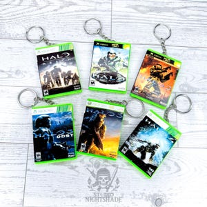 May include: A collection of keyrings showcasing miniature Xbox 360 game cases from the Halo series. Each keyring has a green border and a silver ring. The game covers feature different Halo game titles and characters.