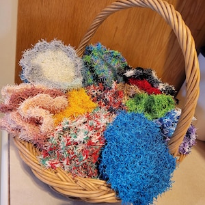 May include: A wicker basket overflowing with colorful, handmade scrubbies. The scrubbies are in various colors, including blue, green, yellow, red, and pink. The basket has a woven handle and is filled with a variety of textures.