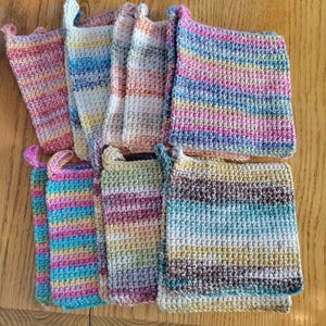 May include: A collection of handmade crochet pot holders in various colors. Each pot holder features a loop for hanging and is made with a striped pattern in shades of pink, blue, yellow, and green. The pot holders are arranged on a wooden surface.