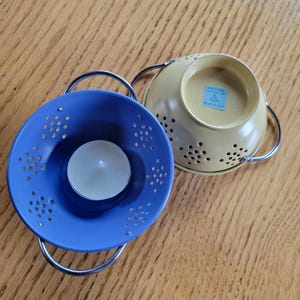 May include: Two small, decorative candle holders. One is blue with a lit tea light inside, the other is yellow. Both have silver handles and a colander design with small holes. The candle holders sit on a wooden surface.