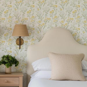 May include: A bedroom scene featuring a cream-colored headboard, white bedding, and a textured beige pillow. A woven lampshade and a small wooden nightstand with a vase of greenery are visible. The wallpaper has a floral pattern with yellow and white flowers.