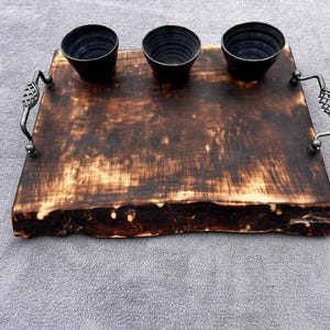 Puede incluir: Una bandeja de madera rústica con tres cuencos negros. La bandeja tiene un borde natural y asas de metal. La madera tiene un acabado oscuro y quemado. Las asas de metal tienen un diseño de jaula decorativo.