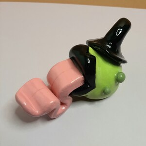 Könnte beinhalten: Eine skurrile Keramikfigur mit grünem Körper, schwarzem Hut und rosa Zunge. Die Figur hat eine glänzende Oberfläche und ein verspieltes Design.