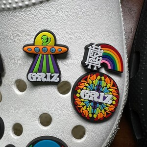 Może przedstawiać: Białe obuwie z kolorowymi zawieszkami Jibbitz, w tym tęczą, UFO z kosmitą i okrągłą zawieszką z napisem "GRIZ" w tęczowym wzorze.
