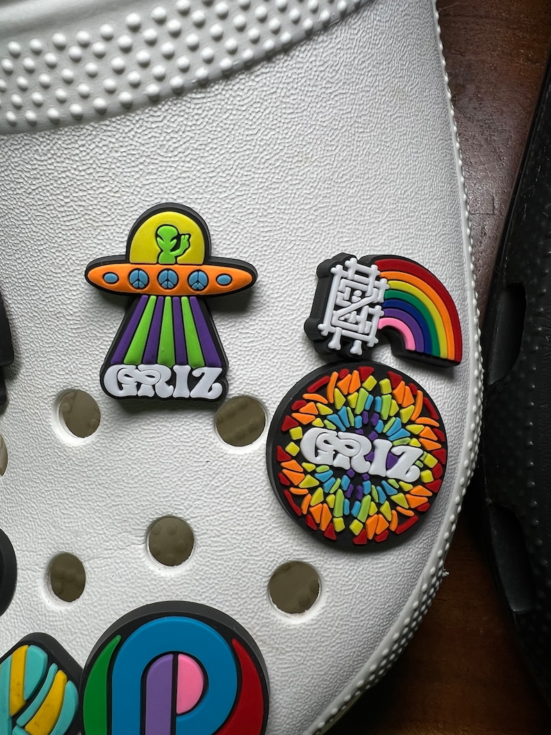 Może przedstawiać: Białe obuwie z kolorowymi zawieszkami Jibbitz, w tym tęczą, UFO z kosmitą i okrągłą zawieszką z napisem "GRIZ" w tęczowym wzorze.