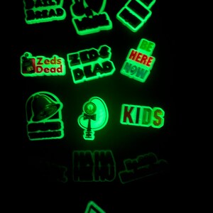 Może przedstawiać: Zbi&oacute;r 12 świecących w ciemności naszywek do prasowania z r&oacute;żnymi wzorami, w tym "Zeds Dead", "Kids", "Daily Bread", "Be Here Now" i znak pokoju z sercem.