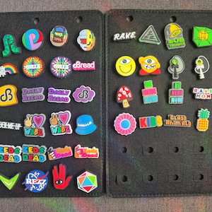 May include: A black felt board with two rows of colorful shoe charms. The charms feature various designs, including cartoon characters, logos, and text. Some of the text includes "Rave Mom", "Kids", "Zeds Dead", "Daily Bread", and "Glass Animals".