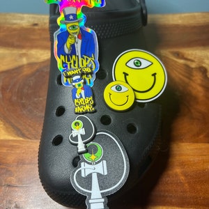 Op de afbeelding: Zwarte klompen met verschillende kleurrijke en grafische schoenbedels. De bedels omvatten een cartoonfiguur met één oog, een smiley met één oog en twee zwart-witte kendama-bedels.
