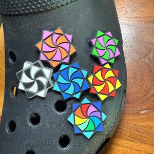 May include: Black clog shoe adorned with colorful, geometric jibbitz charms. The charms feature star-shaped designs with swirling patterns in various colors, including pink, orange, blue, green, yellow, and rainbow hues. The shoe is on a wooden surface.