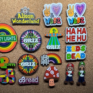 Könnte beinhalten: Eine Sammlung bunter Emaille-Anstecknadeln mit verschiedenen Designs. Die Anstecknadeln enthalten Texte wie "Alison Wonderland", "Pretty Lights", "Griz", "Bread" und "Zeds Dead". Andere Anstecknadeln zeigen einen Pilz, einen Regenbogen und Zeichentrickfiguren.