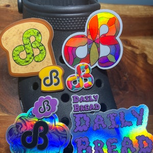May include: A set of colorful and iridescent stickers featuring the letters "DB" in various styles. The stickers are attached to a black shoe with a textured surface. The stickers include a bread-shaped sticker with green swirls, a sticker with a rainbow swirl design, a sticker with a purple cloud design, and a sticker with the text "DAILY BREAD" in purple.