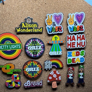 May include: A collection of colorful enamel pins with various designs. The pins feature text like "Pretty Lights," "Alison Wonderland," and "Griz." Other designs include a mushroom, a rainbow, and cartoon characters. The pins are displayed on a corkboard.