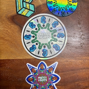 May include: Three colorful round stickers with a geometric design and the text "Pretty Lights" in the center. The stickers are on a wooden surface.