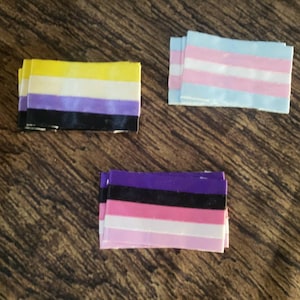 以下が含まれることがあります： 3つの布製の旗で、それぞれ異なる配色になっています。最初の旗は黄色、白、紫、黒です。2番目の旗は水色、白、ピンクです。3番目の旗は紫、黒、ピンク、白です。