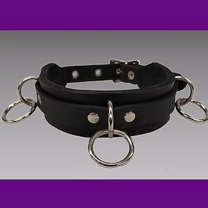 Puede incluir: Choker de cuero negro con tres anillos de metal plateado. El choker es ancho y tiene un acabado liso y pulido.