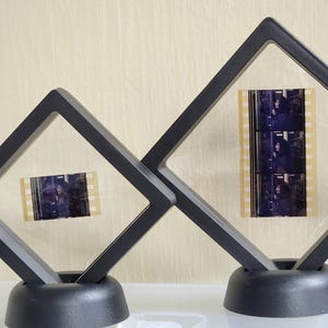 May include: Two black diamond-shaped frames with clear plastic inserts, each holding a strip of film. The frames are on black bases and are sitting on a white surface.