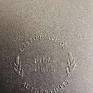 May include: Close-up of a dark grey surface with the words "CERTIFICATE OF AUTHENTICITY" embossed in a circular design. The words "FILM CELL" are in the centre.