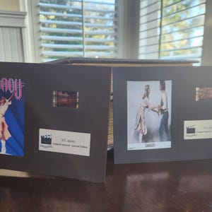 May include: Two black display cases with film strips and movie posters. One case features the word "XANADU" in pink and a woman in a white outfit. The other case has a photo of two people and the word "XANADU". Both cases have a label that says "35 mm Original Insert Special Edition".